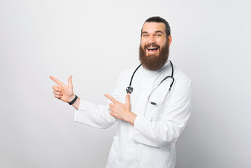 Portrait of cheerful bearded doctor man pointing away at copyspace over white wall