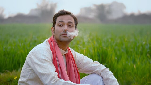 Young North Indian farmer smoking Bidi or Beedi in his green agricultural farm. Medium shot of a handsome village laborer taking a smoke break during his hard work - farmer's leisure time 