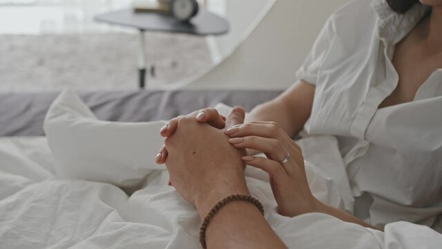 Close-up of unrecognizable romantic couple chilling in bed under white blanket holding and patting each other hands