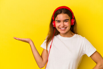 Young caucasian woman listening to music isolated on yellow background showing a copy space on a palm and holding another hand on waist.