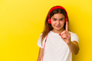 Young caucasian woman listening to music isolated on yellow background showing number one with finger.