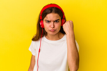 Young caucasian woman listening to music isolated on yellow background showing fist to camera, aggressive facial expression.