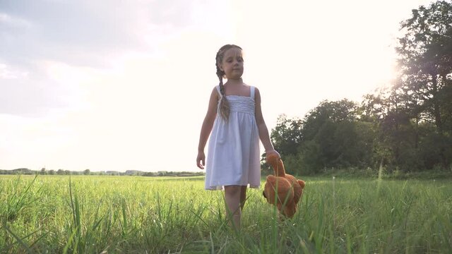 Little girl with a toy teddy bear in park. Cute girl with a teddy bear in a green field. Lonely girl with a toy teddy bear in park in green field.Teddy bear in the hands of a girl in the park