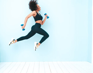 Fototapeta na wymiar Fitness smiling black woman in sports clothing with afro curls hairstyle.She running and jogging . Young beautiful model with perfect tanned body.Female jumping with dumbbells in studio near blue wall