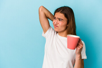 Young caucasian woman holding a mug isolated on blue background touching back of head, thinking and making a choice.