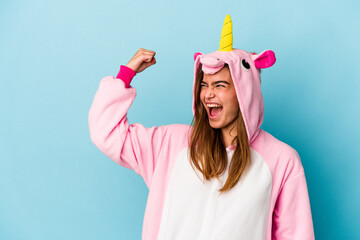 Young caucasian woman wearing a unicorn pajama isolated on blue background raising fist after a victory, winner concept. © Asier