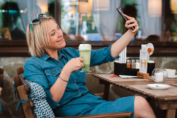 Young woman sitting at a table summer cafe, drinking green matcha drink and doing selfie
