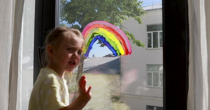 Preschooler girl stands by large window with painted colourful rainbow and waves goodbye against city building and tree closeup