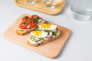 Toasted sandwiches white bread with cream cheese, vegetables: cherry tomatoes, cucumbers, eggs, parsley. Healthy vegetarian breakfast idea. Served with fresh lemon water, light background, copy space