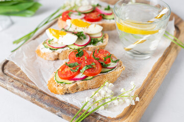 Toasted sandwiches white bread with cream cheese, vegetables: cherry tomatoes, cucumbers, eggs, parsley. Healthy vegetarian breakfast idea. Served with fresh lemon water, light background, copy space