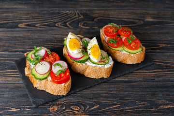 Healthy vegetarian breakfast. Toasted sandwiches white bread with cream cheese, vegetables: cherry tomatoes, cucumbers, eggs, parsley. Served on black stone board, dark wooden background