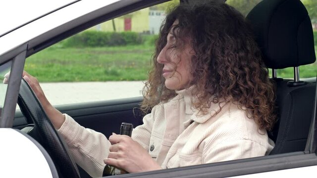 Beautiful middle-aged woman drinks wine from a bottle sitting in her car. She is already very drunk.