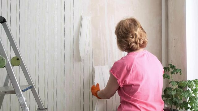 Woman removing old wallpaper from walls preparing for flat renovation. A female hand strips wallpaper from the wall of a room. Room renovations at house. Interior design and home renovation concept