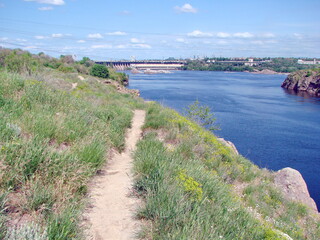 Walk on a narrow path on top of the Dnieper bank on the background of a dam under white clouds on the horizon.