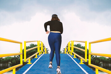 A young woman runs up a hill on a pedestrian bridge over a canal, a blue asphalt road