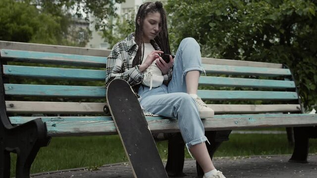 Cool skateboarder girl with braids and nose piercing sitting on bench in city park while texting on her cell phone