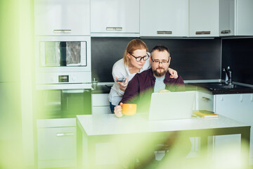 Adult married couple in cozy home interior. Husband and wife look at the laptop screen, discussing family budget. Woman embraces man from behind. In the foreground leaves of houseplant, blurred focus.