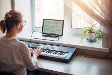 Obraz na płótnie Canvas View from the back young woman plays a synthesizer, reading notes on a laptop screen. Independent learning to play the piano at home. Passion for music, hobbies, leisure, self-development.