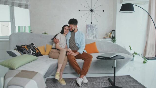 Full-shot portrait of young loving Latin couple sitting on comfortable grey sofa with colorful pillows in modern design living room hugging each other and smiling to camera