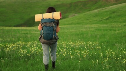 Naklejka na ściany i meble An active young caucasian woman with backpack goes on hike. Girl Traveler travels on green grass and flowers, meditation, ecotourism, hiking. Free woman traveler walking through the meadow in summer.