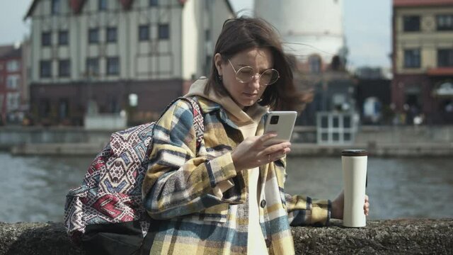 Portrait of young female torist with backpack texting on her cellphone while drinks coffee on enbankment