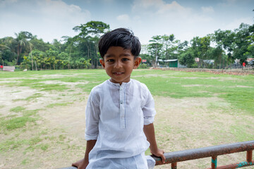 A white Punjabi-wearing smiling Asian kid is sitting on a steel pipe. A Muslim boy is wearing a white Punjabi modern outfit. - obrazy, fototapety, plakaty