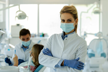 Female dentist posing in modern dental office with crossed arms, protective mask and gloves.