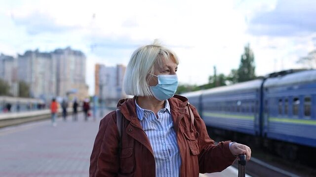 Adult blonde woman in protective medical mask on public train station platform during coronavirus Covid-19 pandemic. Travel by train