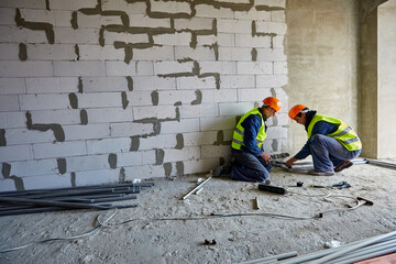 Two males builders in working clothes and safety hard hats are installing plastic pipes using...