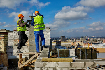 Two males professional workers in protective clothing and hardhats are working with ventilation system on roof of building under construction