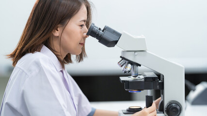 Fototapeta premium A young scientist woman in a laboratory coat looking through a microscope in a laboratory to do research and experiment. Scientist working in a laboratory. Education stock photo