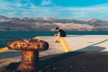 pensive teenager sitting on a pier by the sea with beautiful scenery in the background