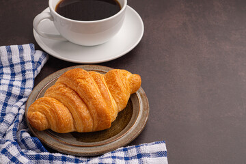 Top view of a croissant on a plate and a white coffee cup with a cloth on a dark gray background. Space for text. Dessert and relaxation concept