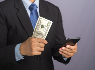 Hand of a businessman using a smartphone and holding US dollar banknotes while standing with a gray background. Space for text. Close-up photo. Business and finance concept