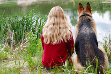 A woman with a dog sits on the shore of the lake. German shepherd and blonde girl for a walk. View...
