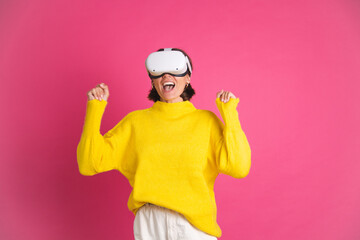 Beautiful woman in bright yellow sweater on pink background in virtual reality glasses happy jumping clenching fist winner gesture