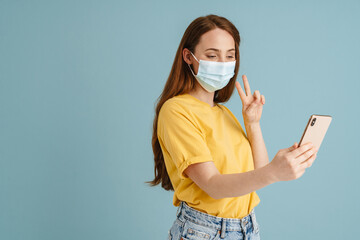 Ginger woman in face mask taking selfie on cellphone and gesturing