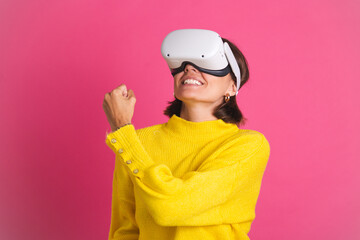 Beautiful woman in bright yellow sweater on pink background in virtual reality glasses happy jumping clenching fist winner gesture