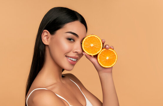 Portrait of Asian girl with shining clean skin of face holding orange halves in white underwear isolated on beige background. Vitamin C cosmetics concept