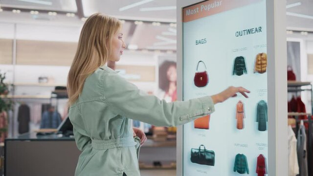 Beautiful Female Customer Using Floor-Standing LCD Touch Display while Shopping in Clothing Store. She is Choosing Stylish Bags, Picking Different Designs from Collection. People in Fashionable Shop.
