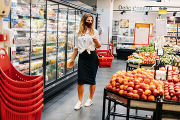 A blonde girl in a protective mask with a grocery basket goes through the store and selects products during the pandemic. Social distance, prevention, isolation