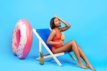Full length of young black lady suffering from summer heat, holding tropical cocktail while sitting...