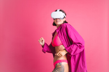 Beautiful fit tanned sporty woman in bikini and shirt in virtual reality glasses on pink background