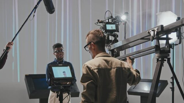 Medium rear-view slowmo of male operator moving camera crane while filming African-American man speaking on camera while gaffer holding lighting working together in studio