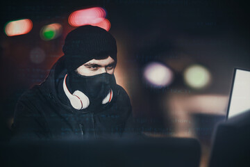 Angry computer hacker weared balaclava stealing data from laptop in front of black background and...