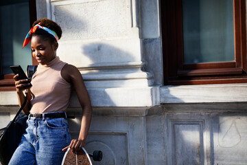 Portrait of happy african-american woman with skateboard. Young stylish woman using the phone outdoors