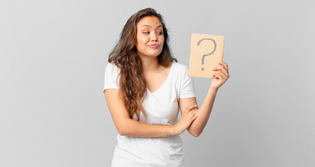 young pretty woman shrugging, feeling confused and uncertain and holding a question mark sign
