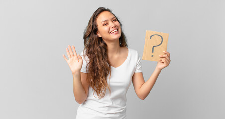 young pretty woman smiling happily, waving hand, welcoming and greeting you and holding a question...