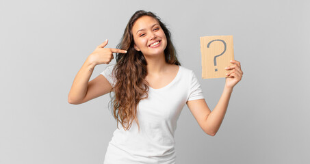 young pretty woman smiling confidently pointing to own broad smile and holding a question mark sign
