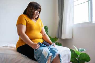 Oversize young woman want to wear short jeans. Overweight female sitting on the bed holding pants...